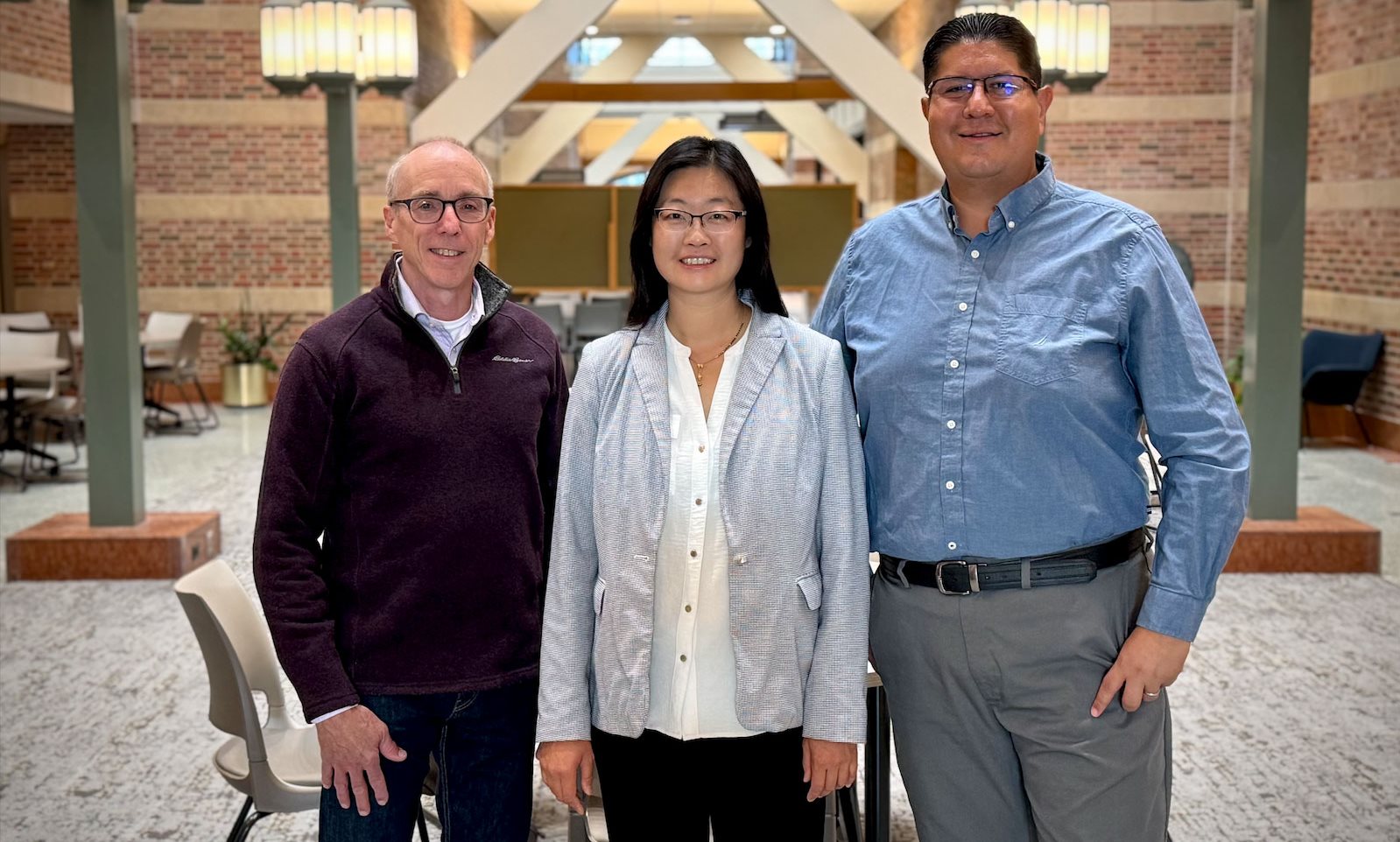a woman with glasses and a light blue jacket standing between a man wearing a maroon sweater and a taller man wearing a blue button down shirt.
