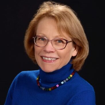 A woman wearing a blue turtleneck and glasses smiling for a professional headshot.