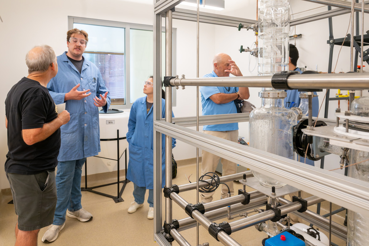 students in lab coats explaining distillation column setup to lab visitors