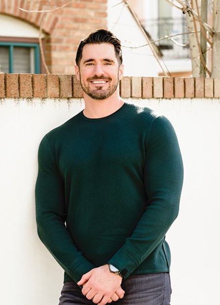 A man wearing a dark green sweater leaning against a wall.