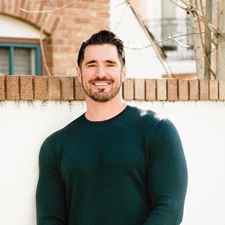Photo shows Adam leaning against a wall, wearing a dark green sweater.