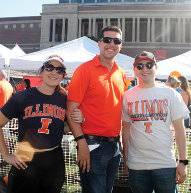 Three smiling alumni wearing orange and blue Illinois attire with the stadium in the background.