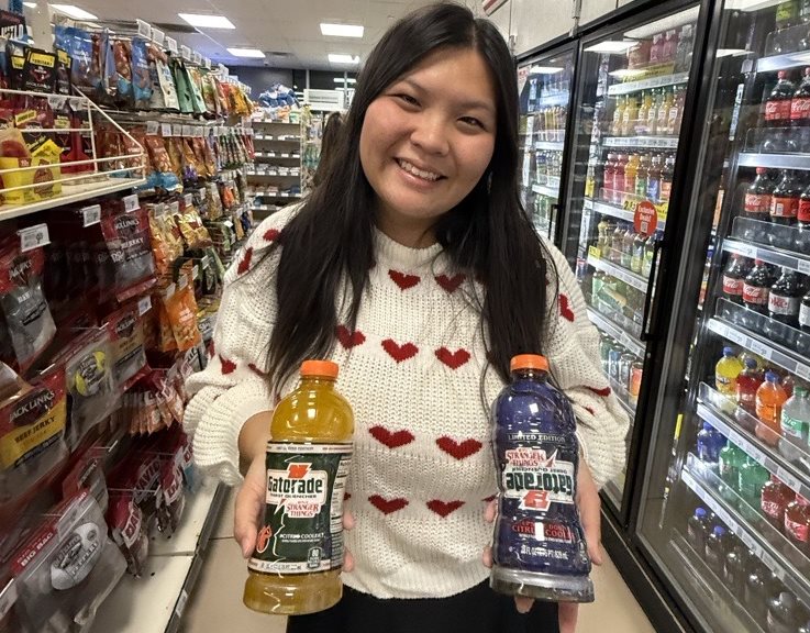 A woman wearing a white sweater with red hearts stands proudly in front of a gas station cooler, holding two flavors of Gatorade.