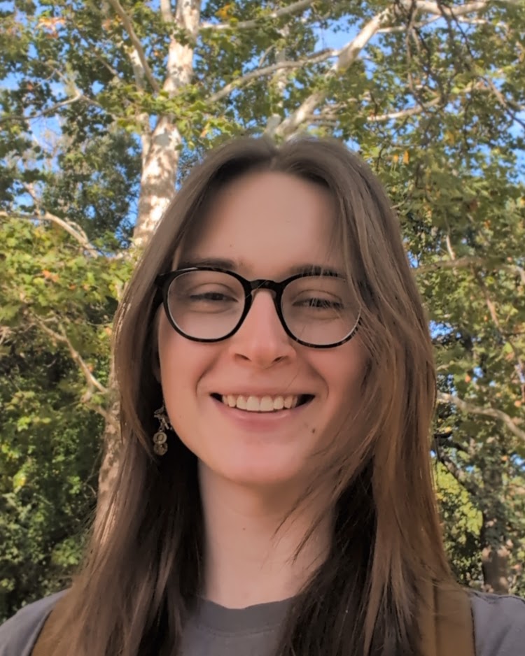 A selfie of a young woman wearing glasses and a gray shirt, surrounded by trees.