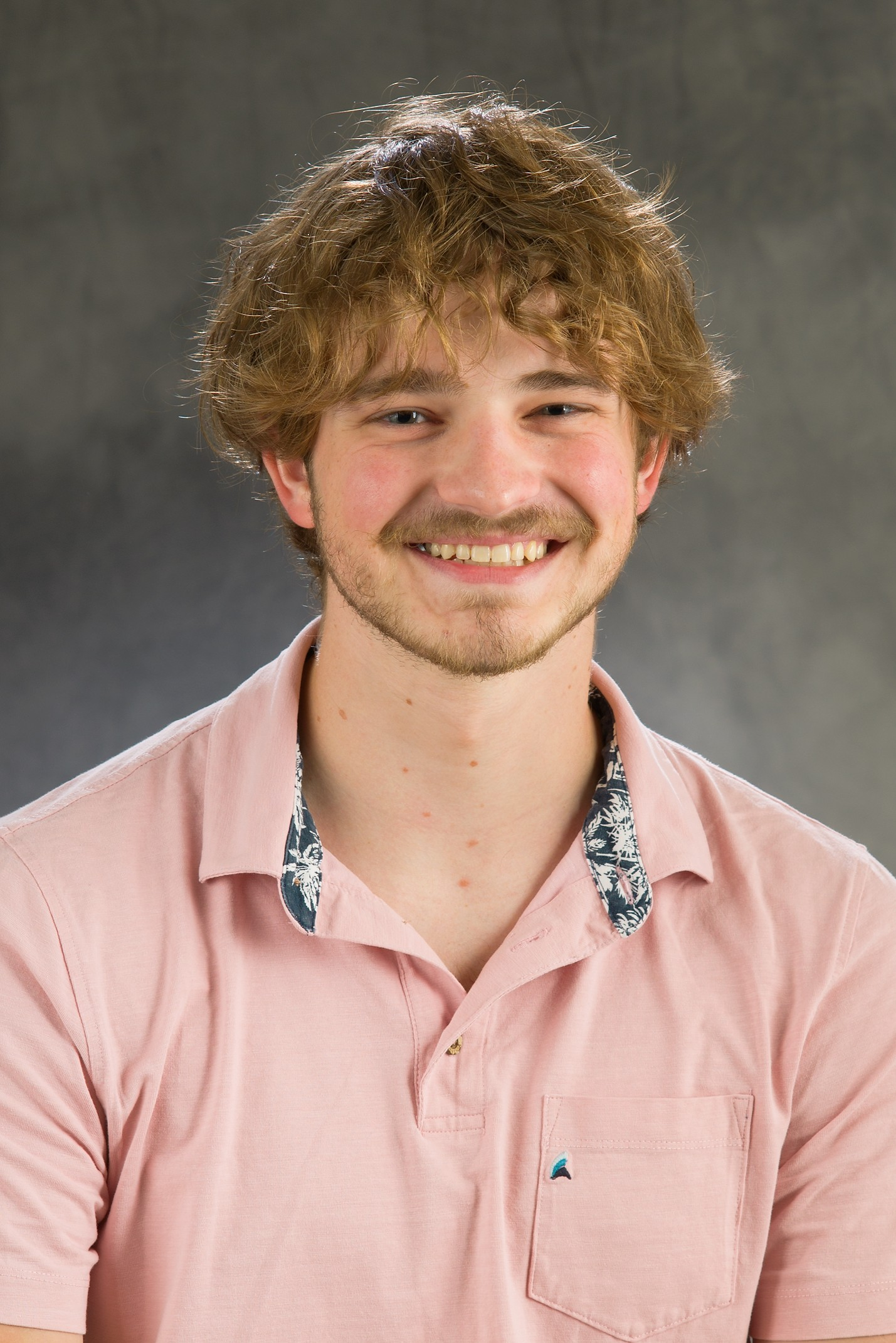 A young man wearing a light pink polo smiles for a professional photo.