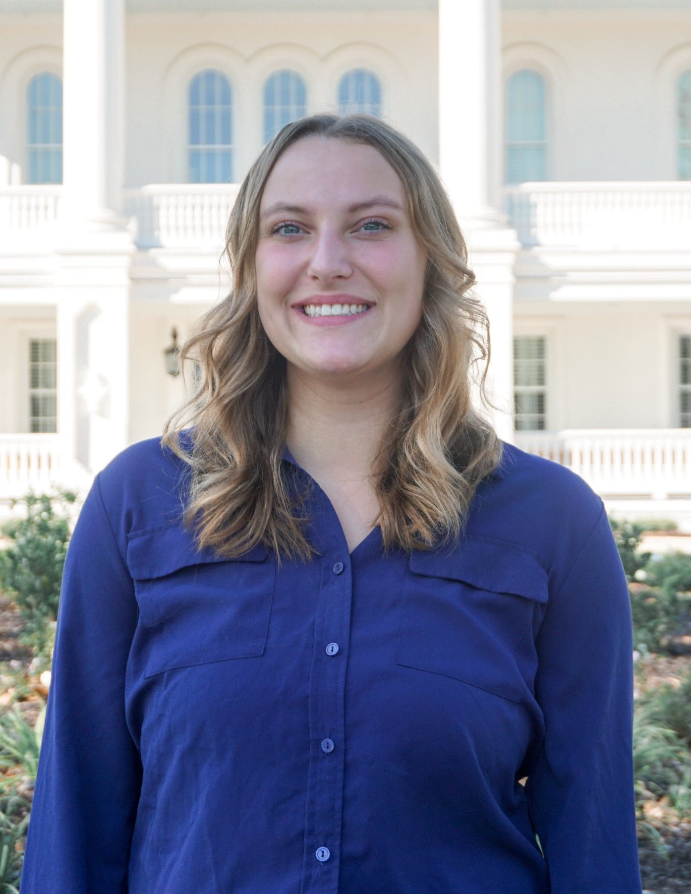 A woman wearing a blue blouse smiles for a professional headshot in front of a grand white building.