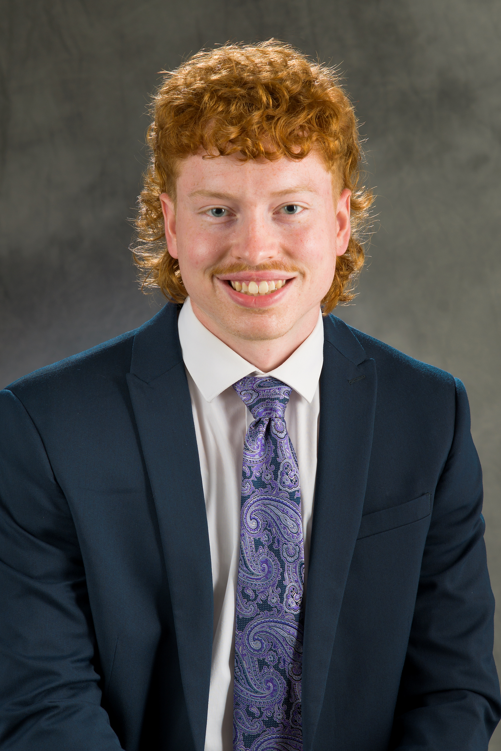 A young man wearing a navy blue suit jacket and patterned purple tie smiles for a professional photo.