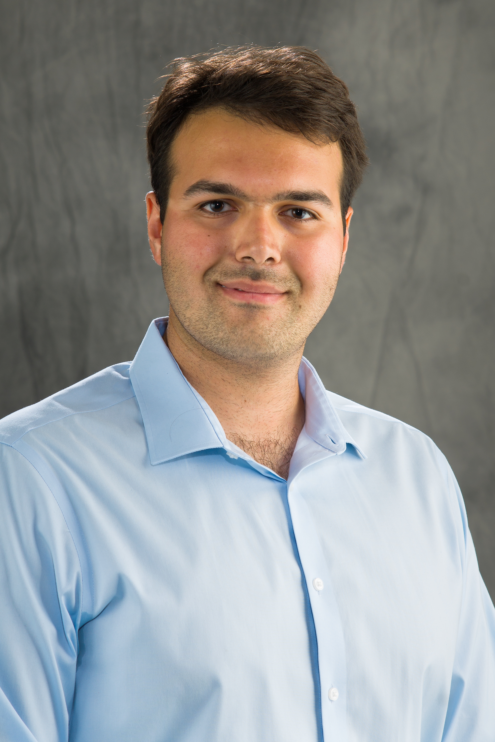 A man wearing a light blue button up smiles for a professional headshot.