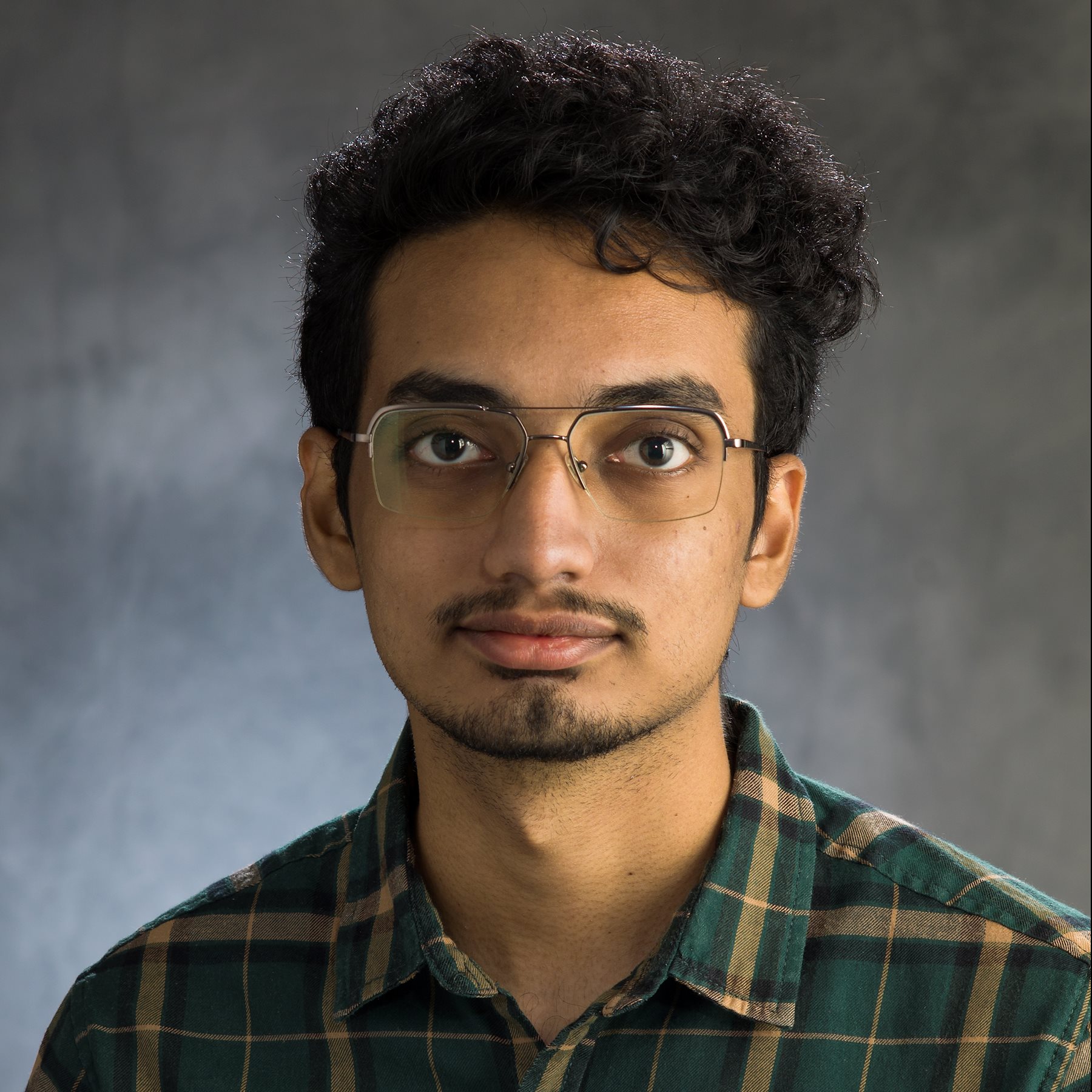A young man wearing glasses and a plaid shirt poses for a professional headshot.