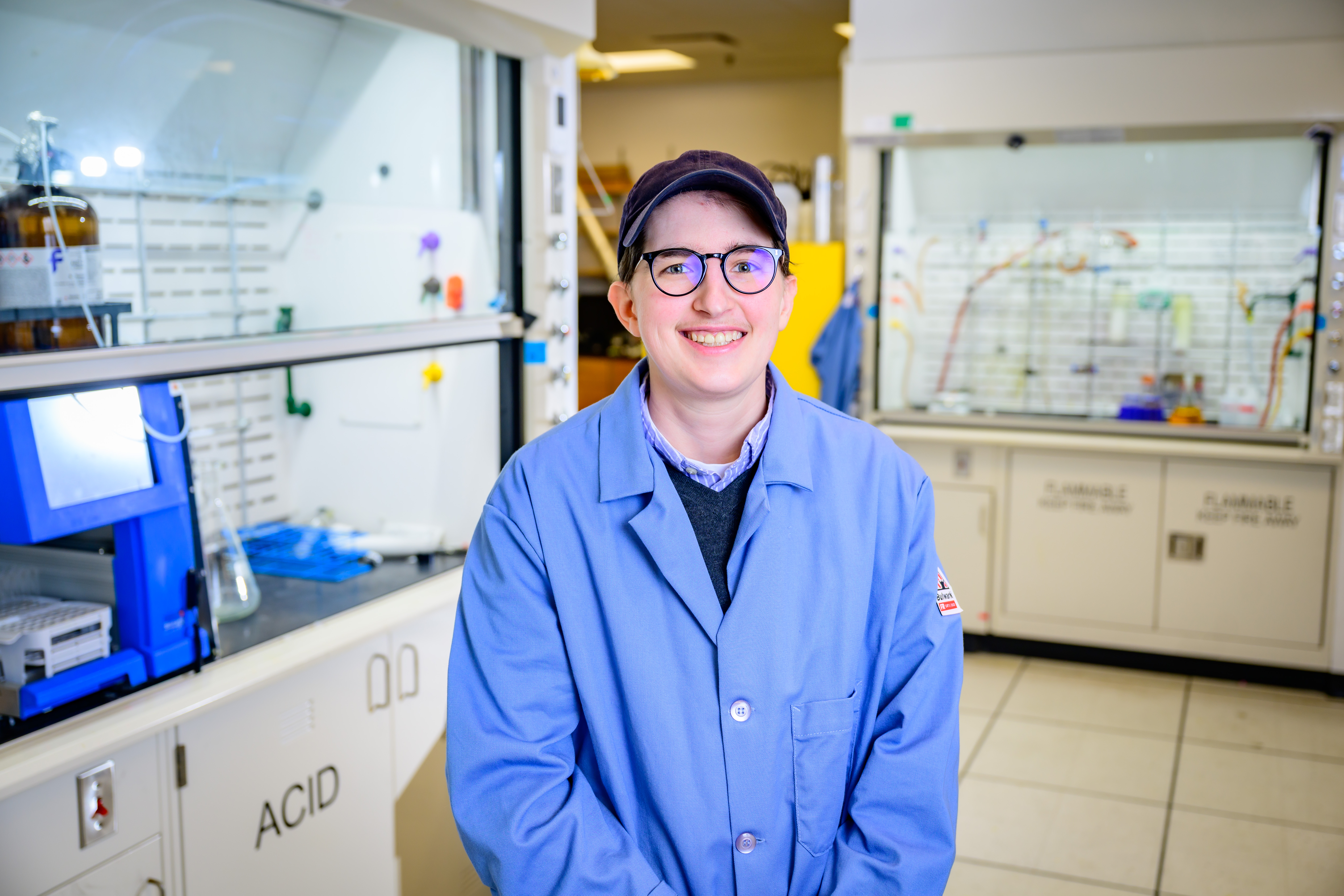 A young woman wearing a blue lab coat, glasses and a baseball cap, smiles proudly in front of her lab equiptment.