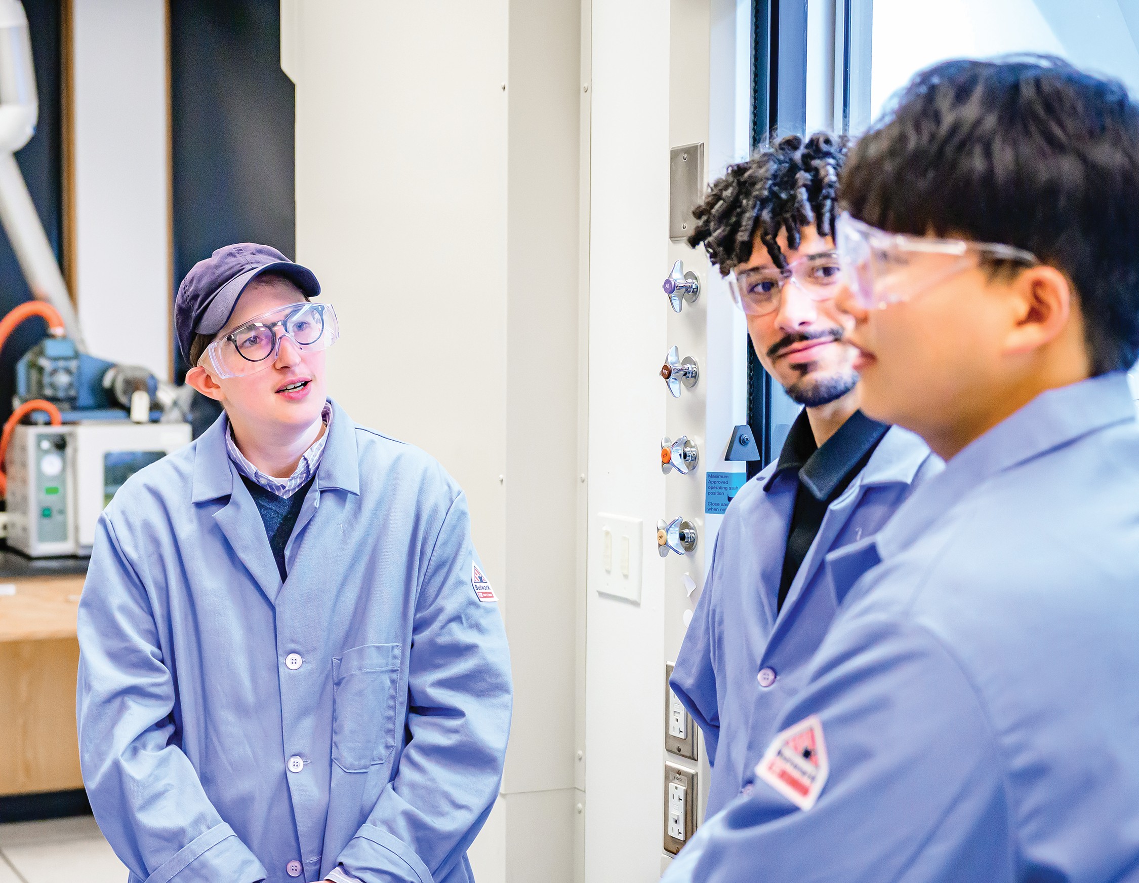 A young woman discusses with her two students, who are wearing protective eye glasses and lab coats.