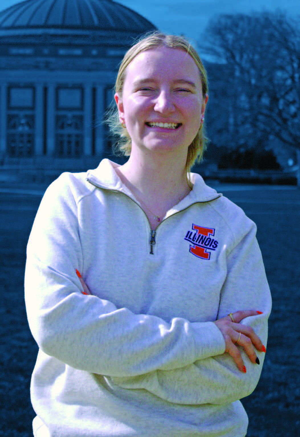 A young woman wearing U of I apparel stands with her arms folded in front of the Quad.