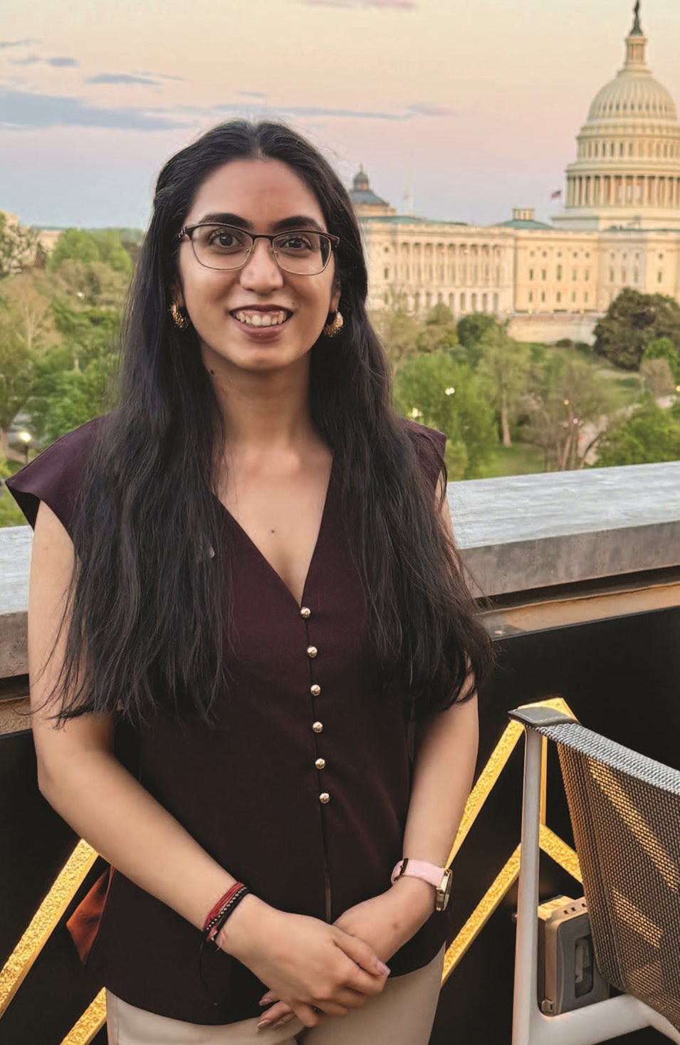 A young woman wearing glasses and a burgundy blouse stands in front of a portion of the US Capitol Building.