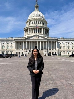 A young woman wearing a black pantsuit stands confidently in front of the United States Capitol Building.