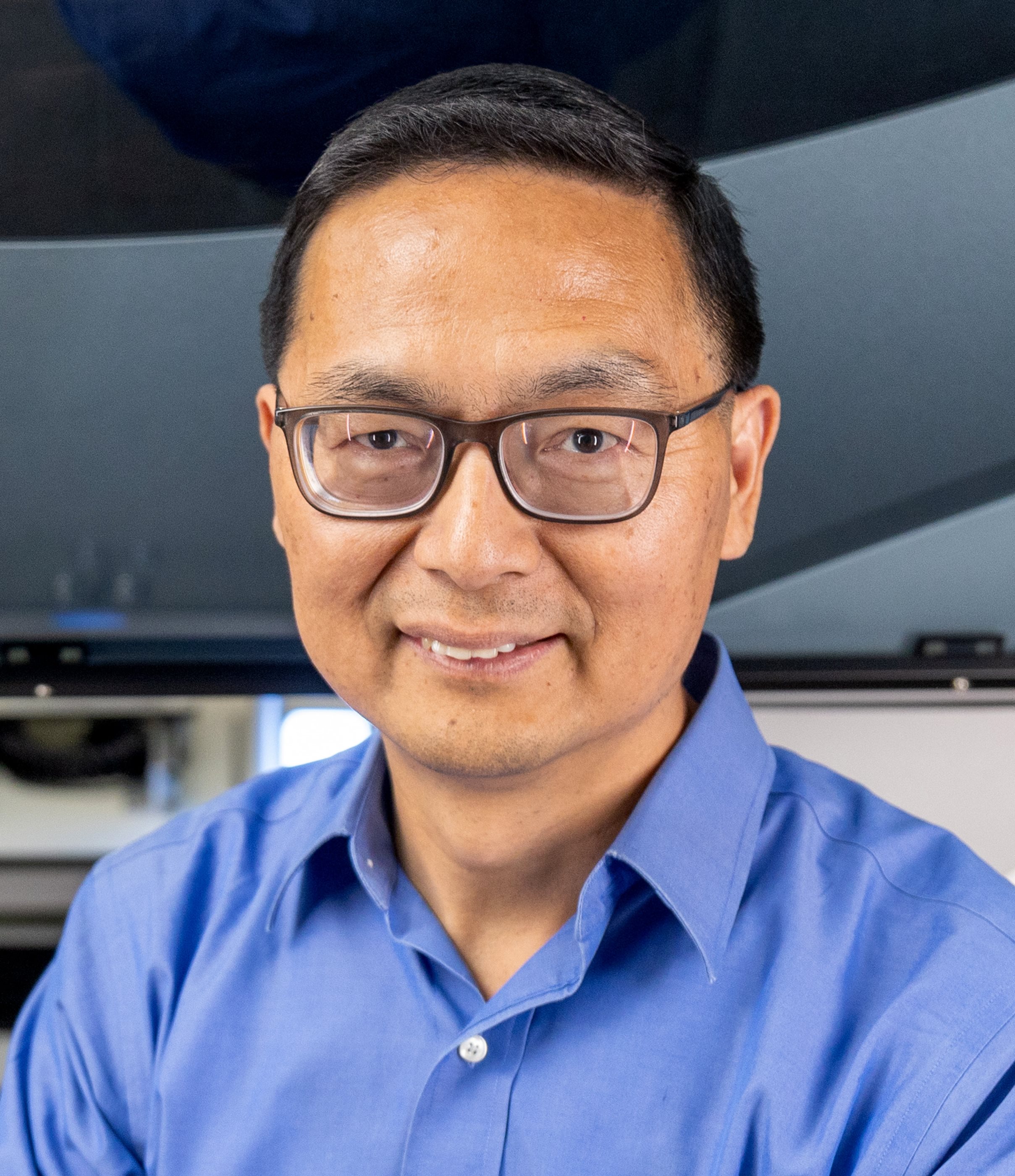 A man wearing a blue button up shirt and glasses smiles for a photo