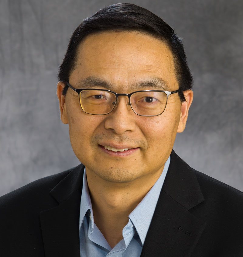A man wearing a black suit jacket and a light blue button up, smiling at the camera.