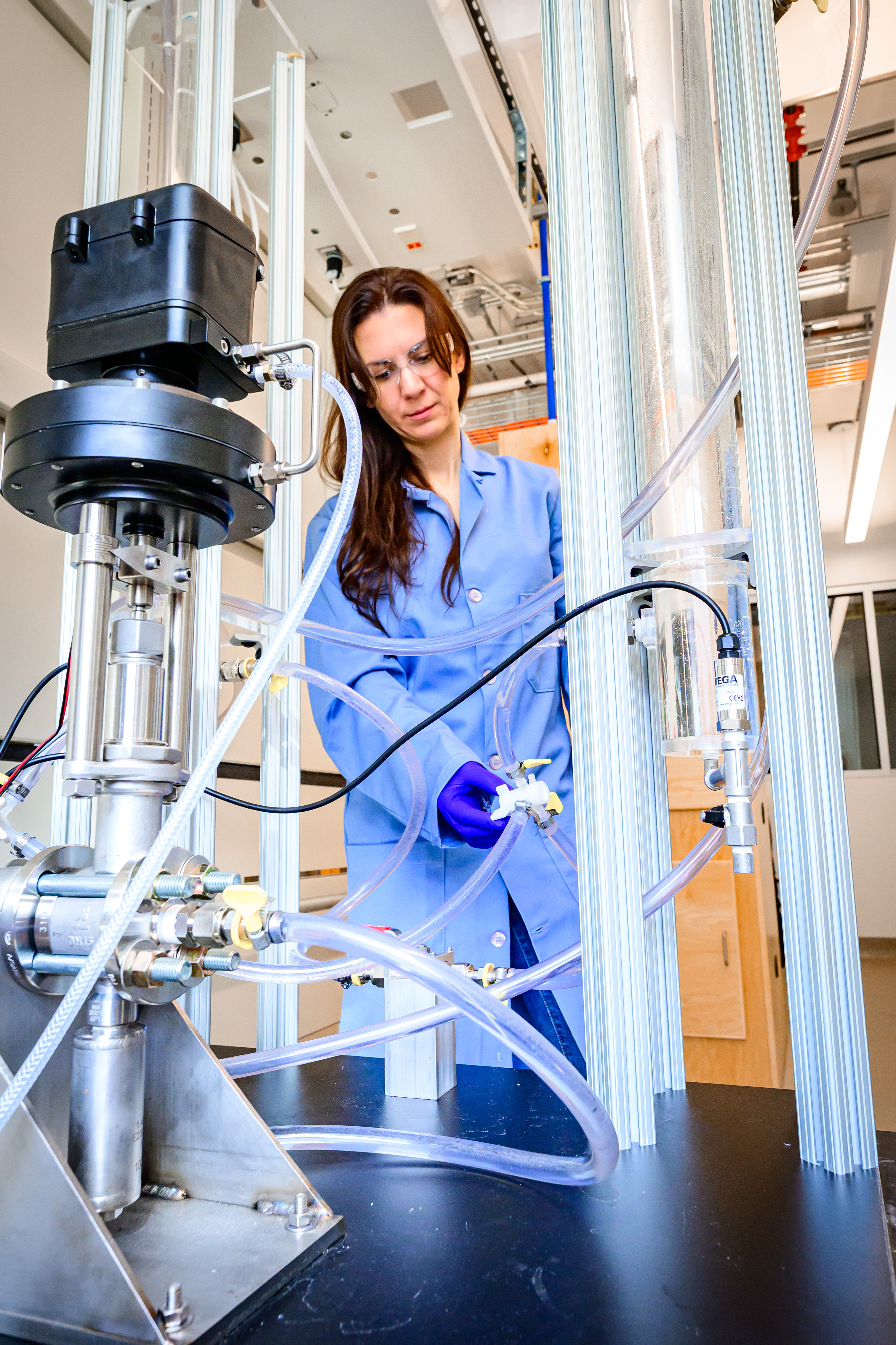 A woman wearing a blue lab coat and protective gear is surrounded by various pumps and test tubes.