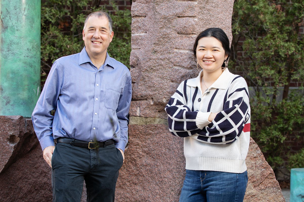 Andrew Gewirth with hands in his pockets and Stephanie Chen with her arms folded lean up against a wall, both smiling at the camera.