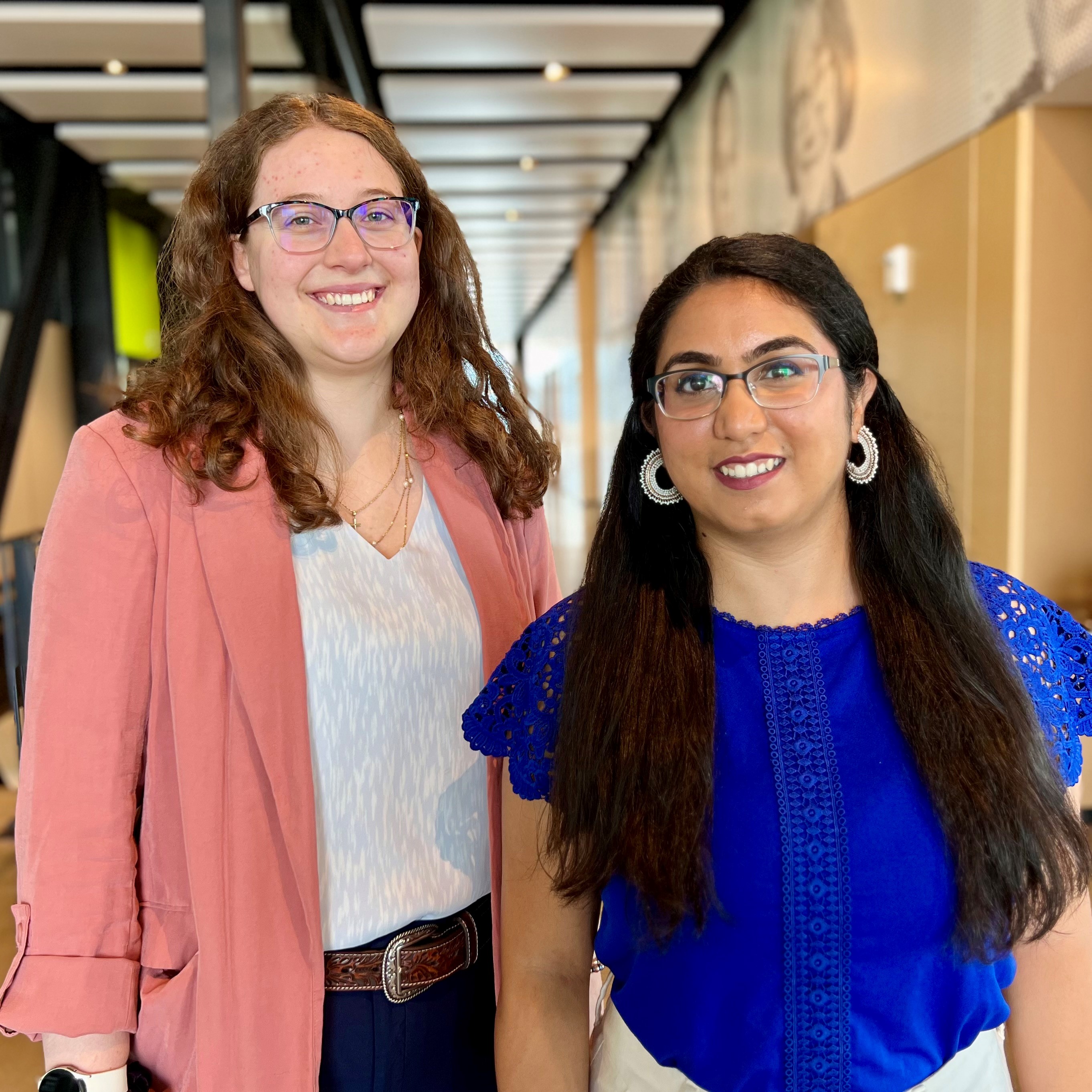 Two women, one with glasses wearing a white blouse and a pink cardigan, and the other also wearing glasses and a blue blouse, stand together, posing for the camera with smiling faces.