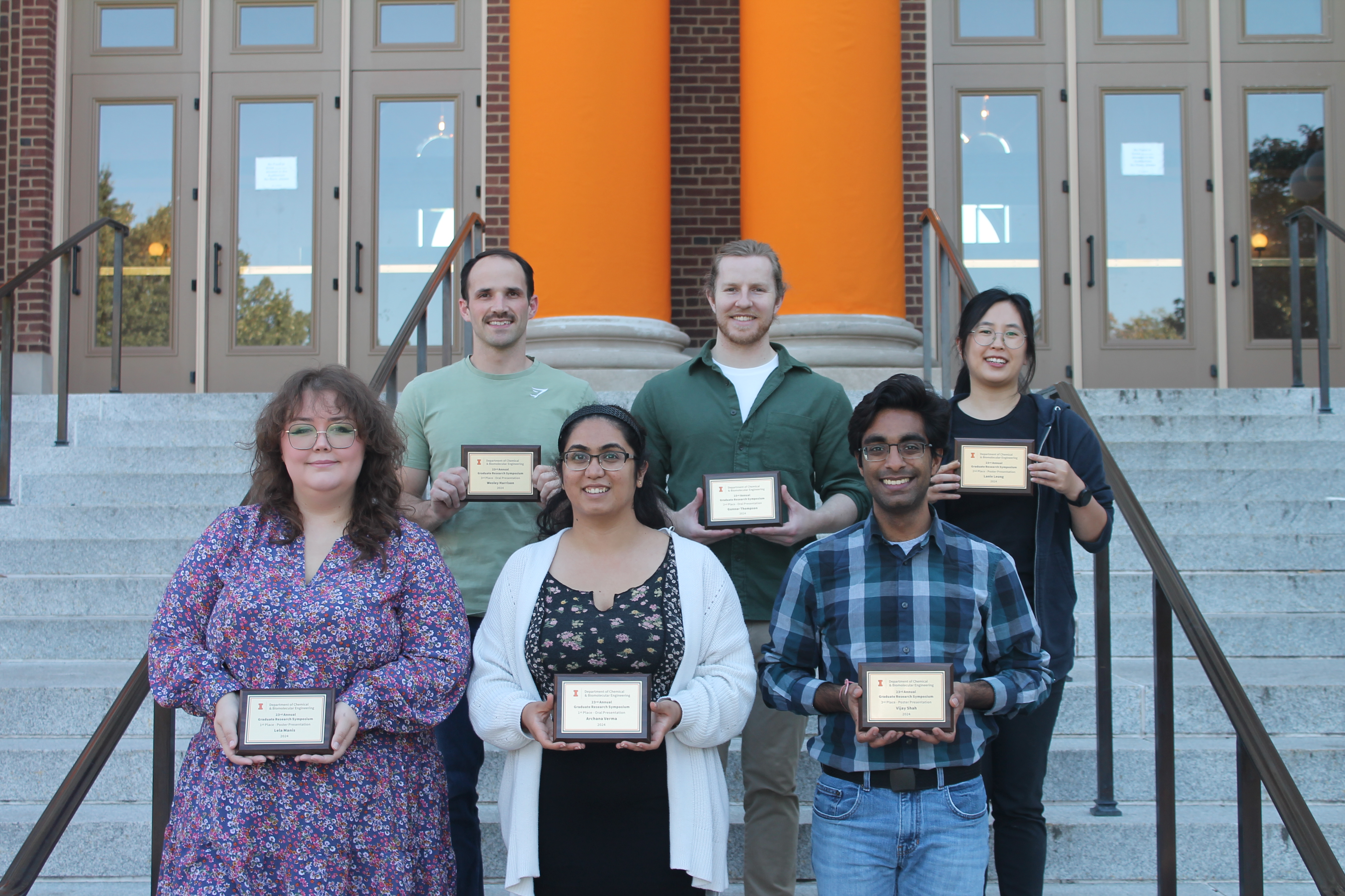 Six people hold up plauqes from winning the Graduate research Symposium on the steps of Follinger Hall.